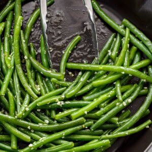 Sautéed, buttery green beans covered in garlic with tongs leaning on side of pan.