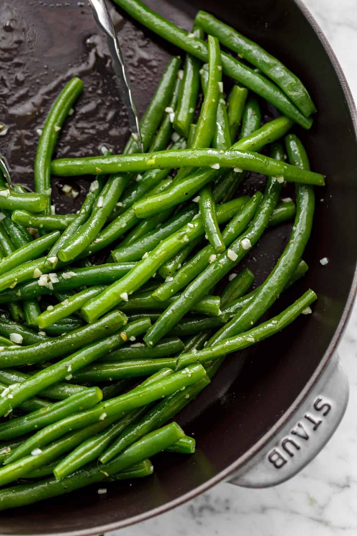 Close up image of Sautéed green beans with a sprinkle of course salt in a pan being pushed aside by a pair of tongs.