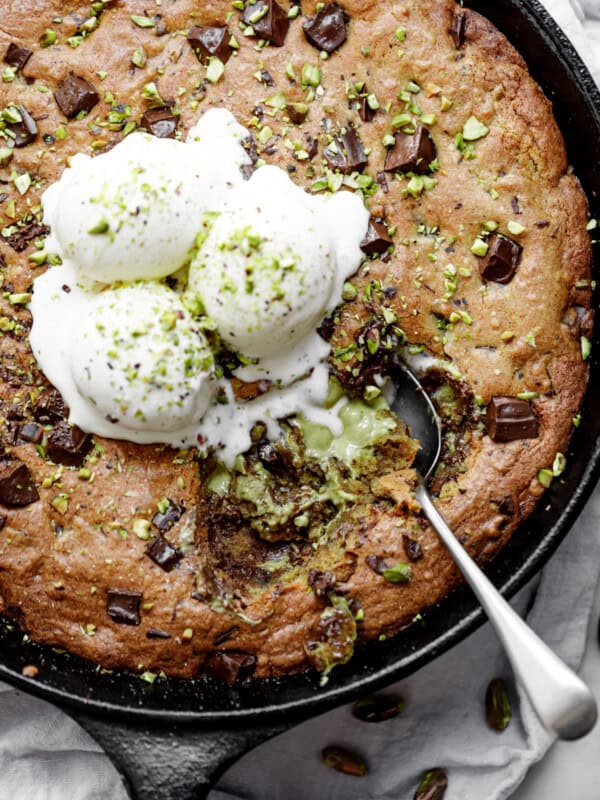 Close-up image of the spoon lifting a portion of pistachio cookie from a skillet, served with vanilla ice cream and topped with chocolate.
